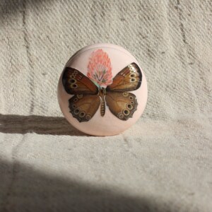 Op de afbeelding: Een ronde, roze sieradenbox met een vlinder- en bloemetjesontwerp. De vlinder is bruin met zwarte markeringen en heeft zijn vleugels uitgespreid. De bloem is roze met een wit centrum.