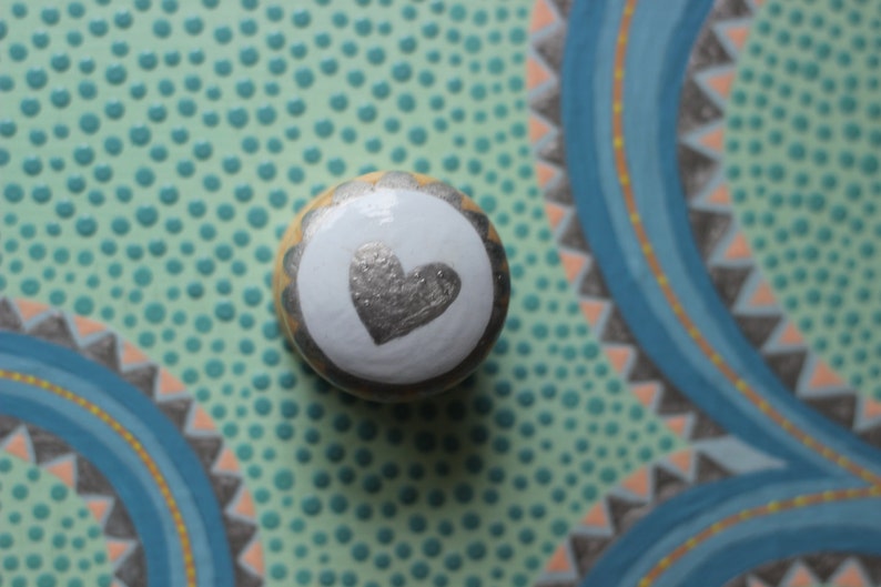 Puede incluir: Un pomo decorativo redondo con un coraz&oacute;n plateado sobre un c&iacute;rculo blanco, rodeado por un borde festoneado dorado y plateado. El pomo est&aacute; sobre una superficie verde claro con dise&ntilde;os pintados en azul y naranja.