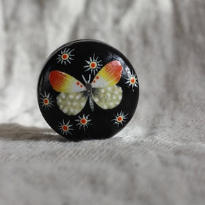 May include: A round, black wooden box with a painted butterfly design. The butterfly has orange, yellow, and white wings with black dots. The box is decorated with white flowers with yellow centres.