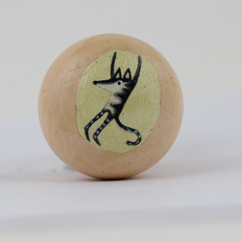 Pode incluir: Objeto decorativo redondo, cor de p&ecirc;ssego, com uma ilustra&ccedil;&atilde;o fantasiosa de uma criatura semelhante a um lobo com os bra&ccedil;os levantados. A ilustra&ccedil;&atilde;o est&aacute; dentro de um oval amarelo p&aacute;lido.