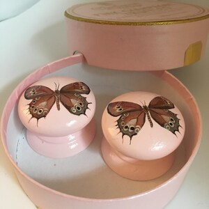 Peut inclure: Deux boutons de placard roses avec un motif de papillon. Les boutons sont ronds et pr&eacute;sentent un papillon peint avec des d&eacute;tails marron et noirs. Ils sont pr&eacute;sent&eacute;s dans une bo&icirc;te rose et ronde avec un couvercle.