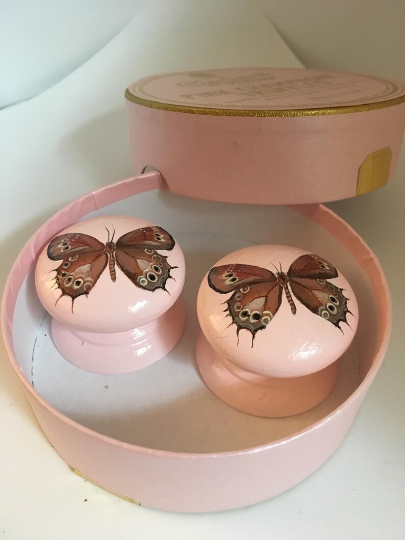 Peut inclure: Deux boutons de placard roses avec un motif de papillon. Les boutons sont ronds et pr&eacute;sentent un papillon peint avec des d&eacute;tails marron et noirs. Ils sont pr&eacute;sent&eacute;s dans une bo&icirc;te rose et ronde avec un couvercle.