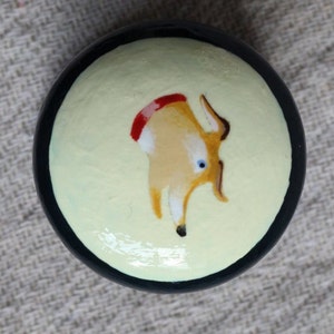 Puede incluir: Un objeto decorativo redondo pintado a mano con un borde negro. El centro presenta un fondo amarillo claro con una ilustraci&oacute;n caprichosa de un ciervo en tonos beige, marr&oacute;n y rojo. El ciervo tiene ojos azules.