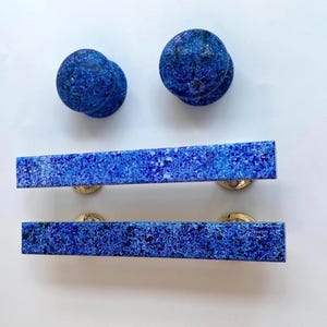 May include: Two blue stone cabinet knobs and two matching rectangular drawer pulls. The hardware has a speckled pattern and is mounted on gold-toned metal bases. The knobs are round and the pulls are rectangular.