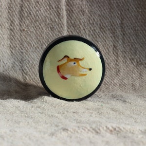 May include: A circular decorative item with a painted dog design. The dog has tan fur, blue eyes, and a red collar, set against a pale yellow background with a black border. The item is likely a decorative element.