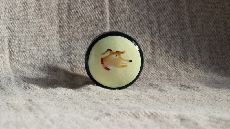 May include: A circular decorative item with a painted dog design. The dog has tan fur, blue eyes, and a red collar, set against a pale yellow background with a black border. The item is likely a decorative element.
