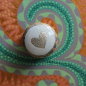 Puede incluir: Un pomo de armario redondo y decorativo con un centro blanco con un dise&ntilde;o de coraz&oacute;n dorado. El pomo se encuentra sobre un fondo colorido con remolinos verdes, grises y naranjas, creando una est&eacute;tica art&iacute;stica vibrante.