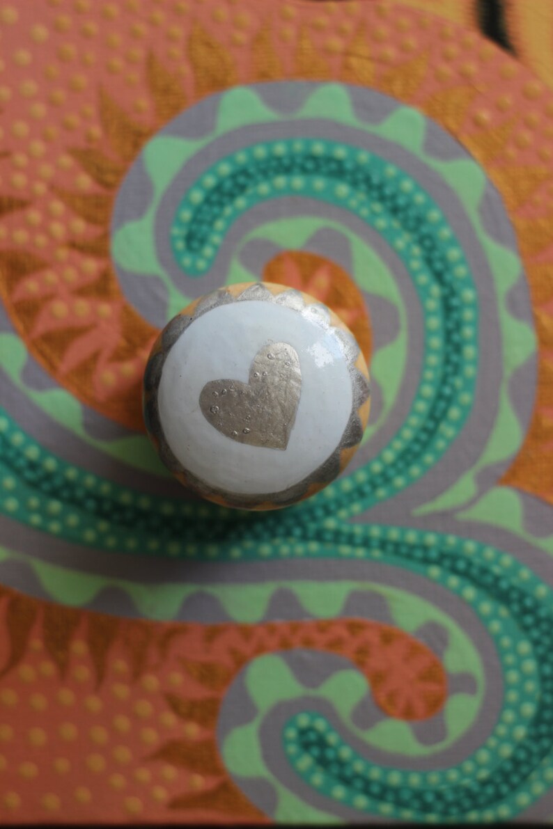 Puede incluir: Un pomo de armario redondo y decorativo con un centro blanco con un dise&ntilde;o de coraz&oacute;n dorado. El pomo se encuentra sobre un fondo colorido con remolinos verdes, grises y naranjas, creando una est&eacute;tica art&iacute;stica vibrante.