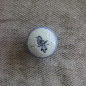 May include: A round, decorative object with a pale blue border and a white centre. A grey bird perched on a thin line is painted in the centre. The object is set against a neutral, textured background.