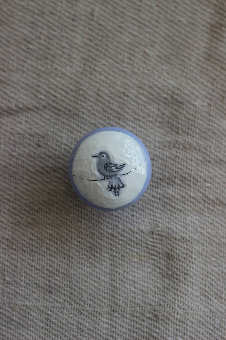 May include: A round, decorative object with a pale blue border and a white centre. A grey bird perched on a thin line is painted in the centre. The object is set against a neutral, textured background.