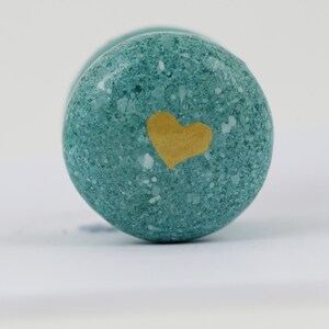 Puede incluir: Primer plano de un objeto redondo de color turquesa con un corazón dorado pintado en la superficie. El objeto tiene una textura moteada y parece ser de piedra. El fondo es blanco liso.