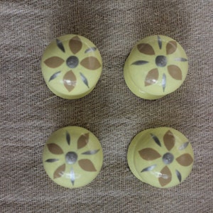 May include: Four yellow ceramic cabinet knobs with a floral design. Each knob features a central grey circle surrounded by brown and silver petal shapes. The knobs are arranged on a textured, neutral-coloured fabric.