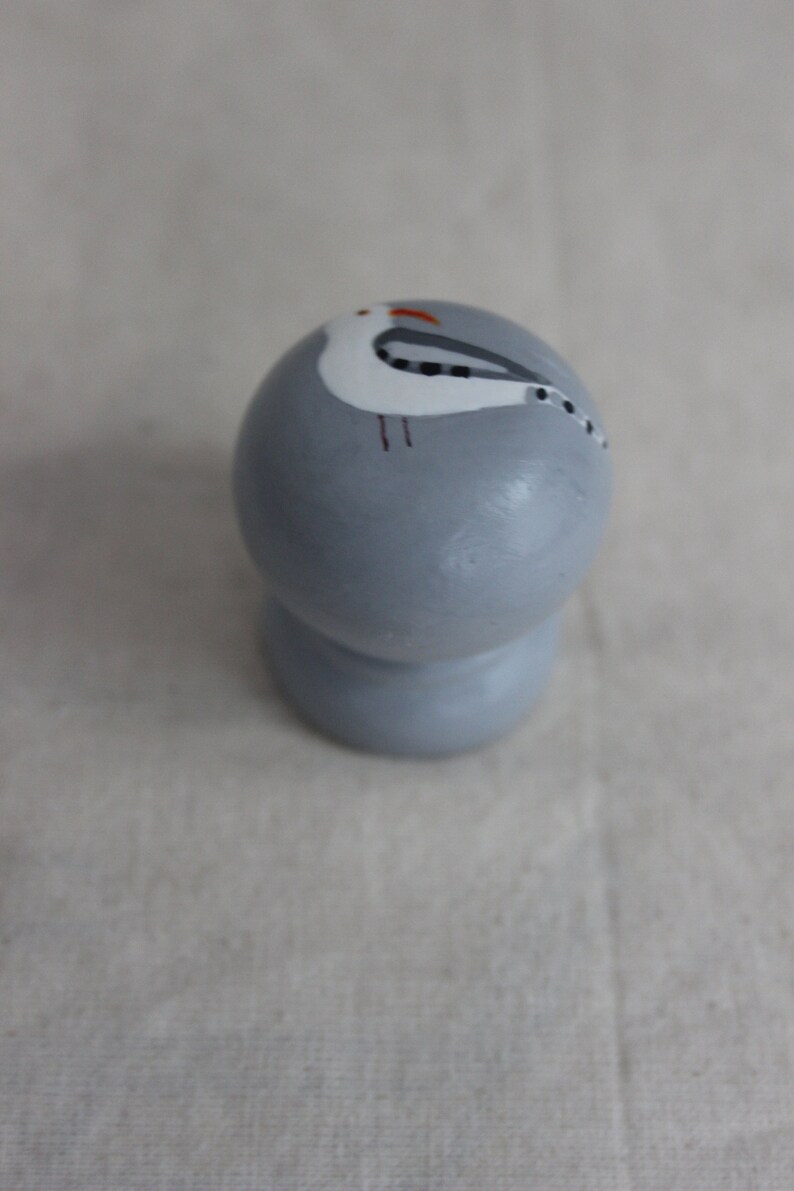 K&ouml;nnte beinhalten: Ein grauer Keramik-Knopf mit einem bemalten wei&szlig;en M&ouml;wen-Design. Die M&ouml;we hat schwarze Augen und einen roten Schnabel.