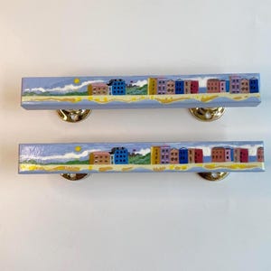 Pode incluir: Duas puxadores de armário azul claro com uma cena costeira pintada. A cena retrata uma fileira de edifícios coloridos ao longo de uma praia com um sol amarelo e ondas brancas. Os puxadores têm ferragens de montagem douradas.