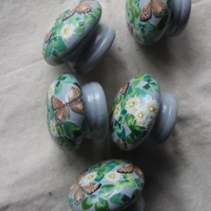 K&ouml;nnte beinhalten: F&uuml;nf graue Keramik-Schrankkn&ouml;pfe mit einem Schmetterlings- und Blumendesign. Die Kn&ouml;pfe sind mit gr&uuml;nen Bl&auml;ttern, wei&szlig;en Bl&uuml;ten und braunen Schmetterlingen bemalt.