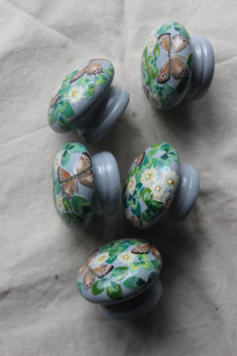 K&ouml;nnte beinhalten: F&uuml;nf graue Keramik-Schrankkn&ouml;pfe mit einem Schmetterlings- und Blumendesign. Die Kn&ouml;pfe sind mit gr&uuml;nen Bl&auml;ttern, wei&szlig;en Bl&uuml;ten und braunen Schmetterlingen bemalt.