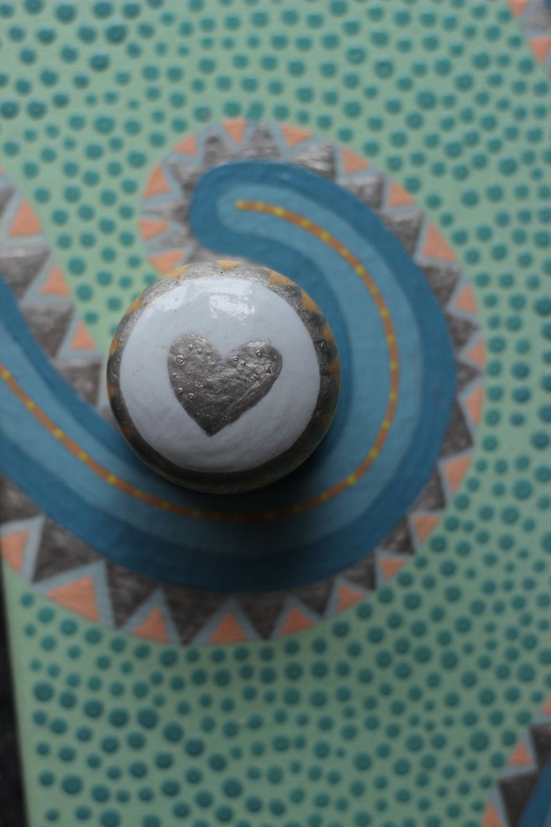 Pu&ograve; includere: Un pomello decorativo per mobili con un centro bianco con un design a cuore argentato. Il pomello &egrave; ambientato su uno sfondo turchese con un vortice blu e motivi geometrici arancioni e grigi. Un accento unico per i mobili.