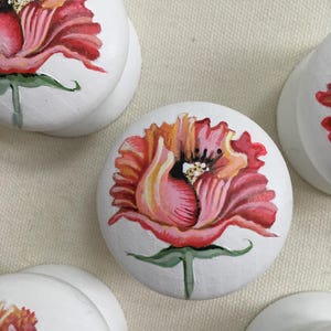 Puede incluir: Pomos de armario redondos blancos con dise&ntilde;os florales pintados a mano. Cada pomo presenta una vibrante flor de amapola roja y naranja con tallos y hojas verdes. Los pomos son un acento decorativo para muebles.