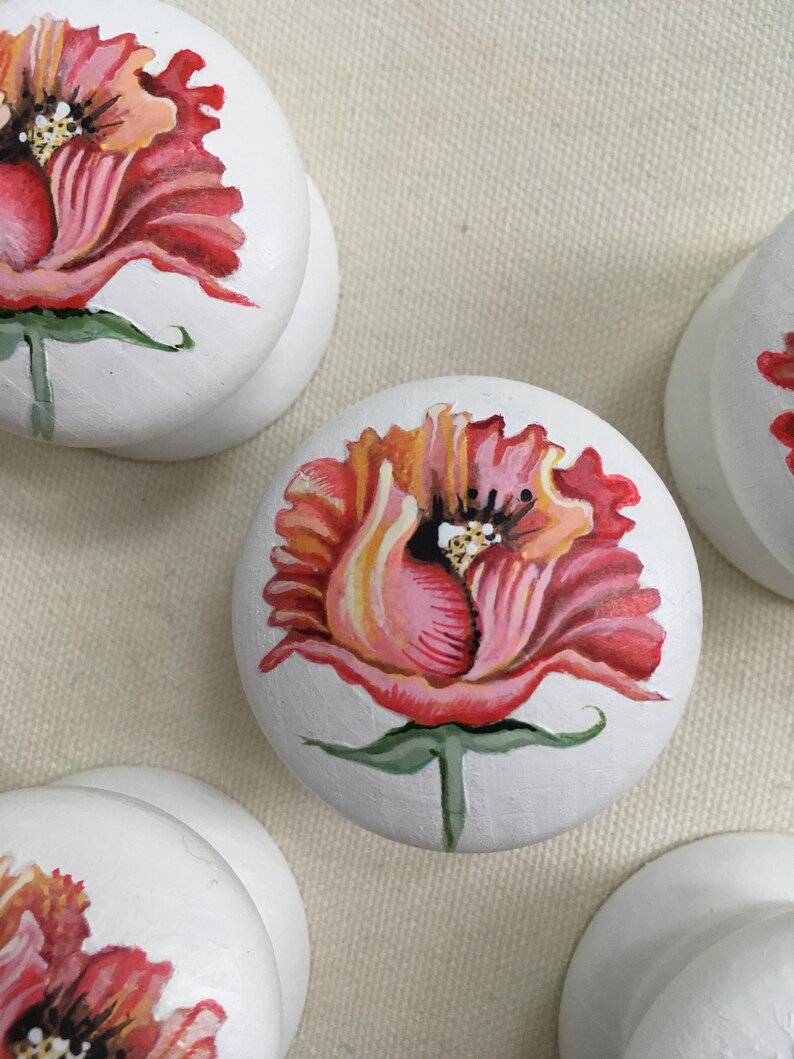Puede incluir: Pomos de armario redondos blancos con dise&ntilde;os florales pintados a mano. Cada pomo presenta una vibrante flor de amapola roja y naranja con tallos y hojas verdes. Los pomos son un acento decorativo para muebles.