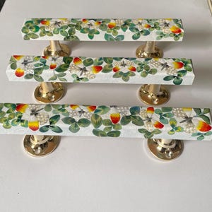 May include: Three decorative cabinet pulls with a white background, featuring a pattern of green clovers, butterflies, and red and yellow accents. The pulls are rectangular with gold-coloured bases.
