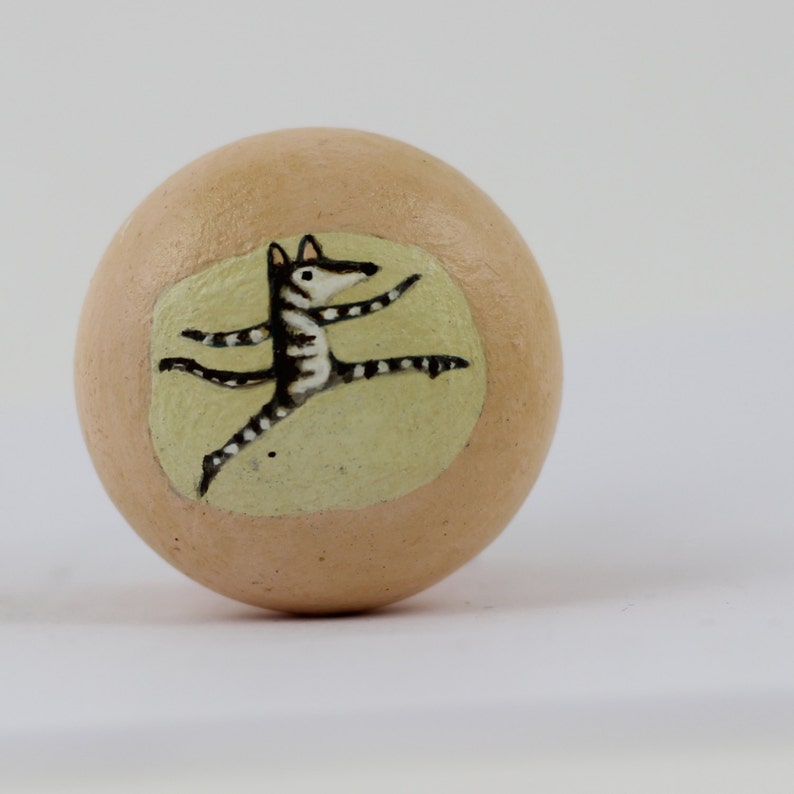 Pode incluir: Objeto decorativo em forma de ovo, bege, com uma ilustra&ccedil;&atilde;o pintada de um animal dan&ccedil;ando. O animal &eacute; preto e branco, com bra&ccedil;os e pernas estendidos, sobre um fundo amarelo p&aacute;lido.