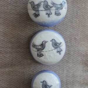 May include: Three circular decorative items with a white surface and a pale blue border. Each features a hand-painted design of grey birds on a wire. The birds have detailed wings and are arranged in different groupings.