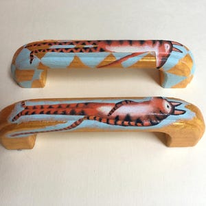 Puede incluir: Dos tiradores de armario de madera con un diseño pintado a mano. Los tiradores son rectangulares con una parte superior e inferior curvadas. Cada tirador presenta un fondo azul claro con una ilustración de gato naranja y dorado.
