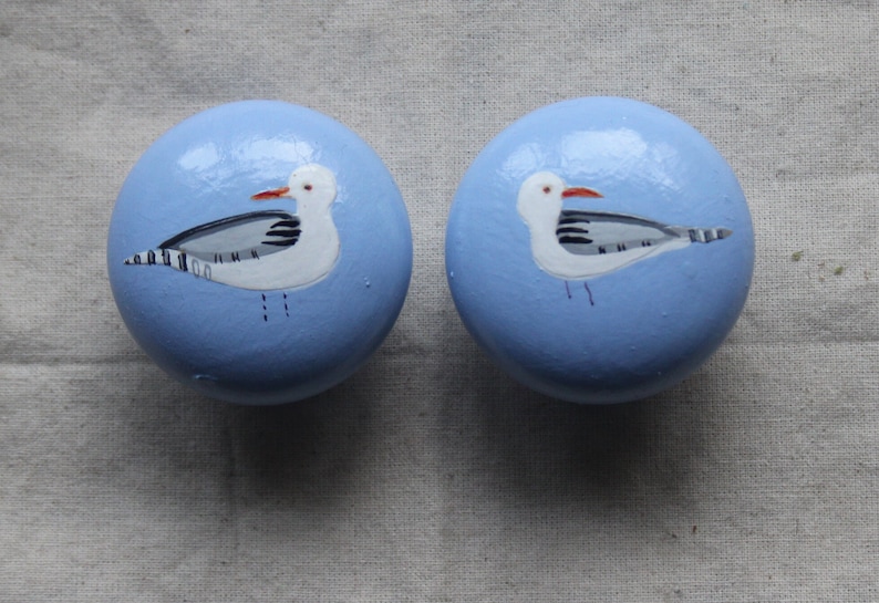 May include: Two light blue round knobs with a white seagull painted on each. The seagulls have black and grey markings and red beaks.