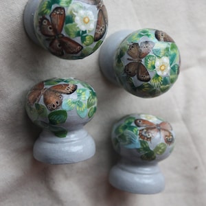 May include: Four round, gray, ceramic knobs with a painted design of butterflies and green leaves. The knobs are painted with a light gray base and have a raised, round base.