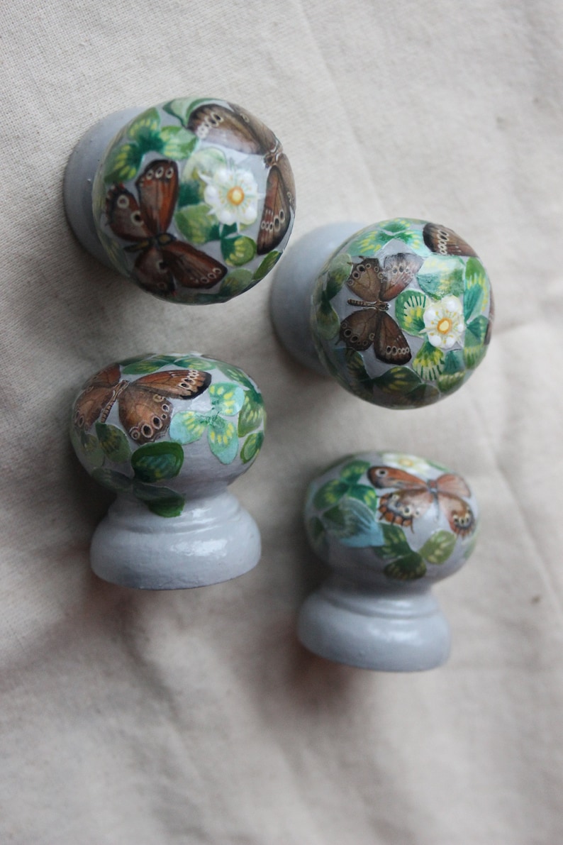 Puede incluir: Cuatro pomos redondos de cer&aacute;mica gris con un dise&ntilde;o pintado de mariposas y hojas verdes. Los pomos est&aacute;n pintados con una base gris claro y tienen una base redonda elevada.