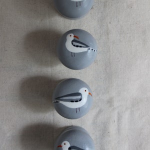 K&ouml;nnte beinhalten: Vier graue runde Keramikkn&ouml;pfe mit bemalten wei&szlig;en M&ouml;wen. Jeder Knopf zeigt eine M&ouml;we mit grauen und wei&szlig;en Fl&uuml;geln und einem orangefarbenen Schnabel.