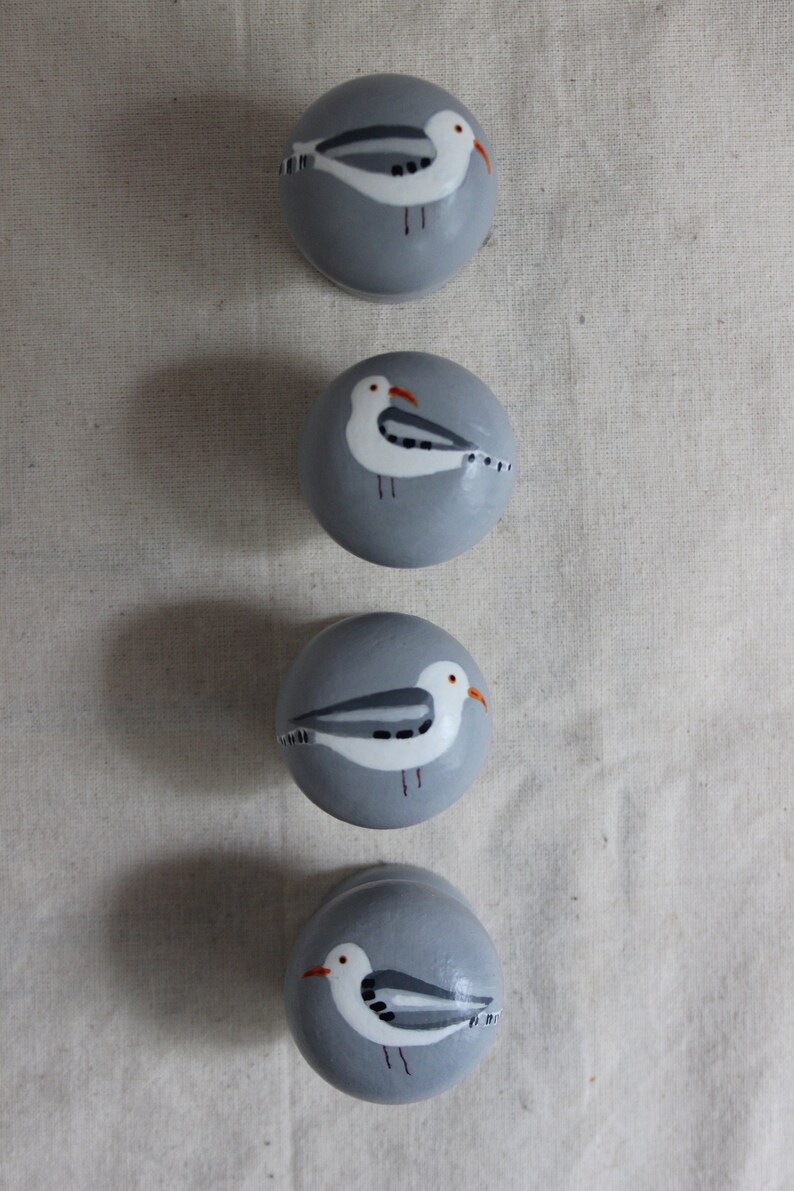K&ouml;nnte beinhalten: Vier graue runde Keramikkn&ouml;pfe mit bemalten wei&szlig;en M&ouml;wen. Jeder Knopf zeigt eine M&ouml;we mit grauen und wei&szlig;en Fl&uuml;geln und einem orangefarbenen Schnabel.