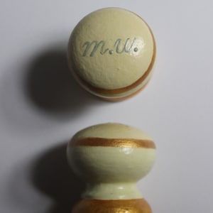 Puede incluir: Dos pomos decorativos de madera. El pomo superior es redondo con las iniciales "m.w." en azul claro. Ambos pomos son de color crema con detalles dorados y marrones. El pomo inferior tiene una base de pedestal.