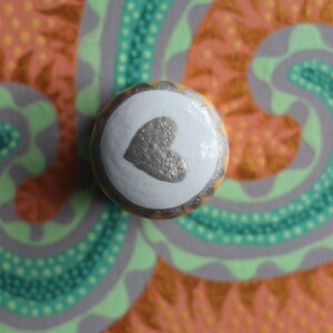 Puede incluir: Pomo decorativo redondo con un coraz&oacute;n plateado sobre fondo blanco, bordeado de oro y marr&oacute;n. El fondo presenta un dise&ntilde;o colorido con tonos naranja, verde y gris.