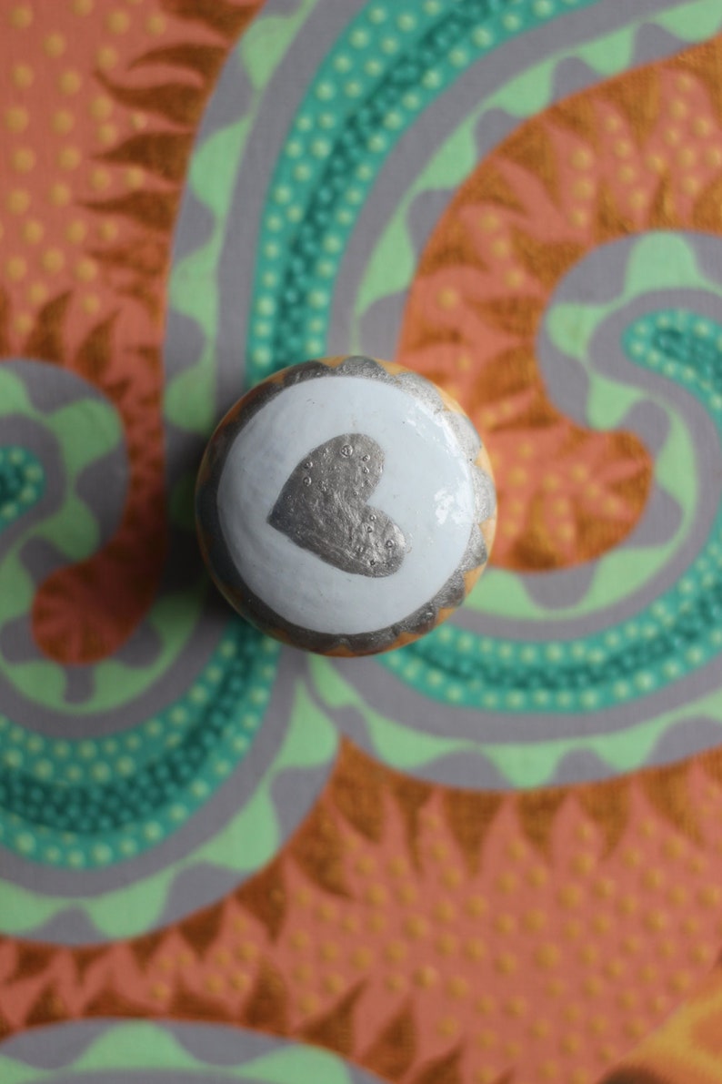 Puede incluir: Pomo decorativo redondo con un coraz&oacute;n plateado sobre fondo blanco, bordeado de oro y marr&oacute;n. El fondo presenta un dise&ntilde;o colorido con tonos naranja, verde y gris.