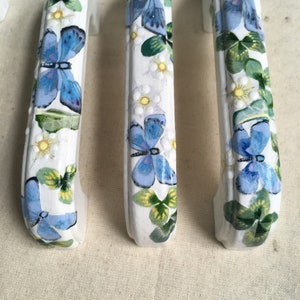 Puede incluir: Tres tiradores de cajón de cerámica blanca con un diseño de mariposa y trébol. Las mariposas están pintadas en tonos azules y los tréboles son verdes.