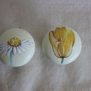 Può includere: Due pomelli rotondi in legno dipinti con motivi floreali. Un pomello presenta una margherita bianca con un centro giallo. L'altro pomello presenta un tulipano giallo.