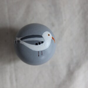 K&ouml;nnte beinhalten: Eine graue bemalte Kugel mit einer darauf gemalten wei&szlig;en M&ouml;we. Die M&ouml;we hat einen grauen R&uuml;cken, einen wei&szlig;en Bauch und einen orangefarbenen Schnabel.