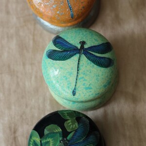 Może przedstawiać: Trzy okrągłe drewniane ozdoby z malowanymi wzorami ważek. G&oacute;rna ozdoba jest pomarańczowa z czerwoną i białą ważką. Środkowa ozdoba jest zielona z niebieską ważką. Dolna ozdoba jest czarna z zieloną i niebieską ważką.