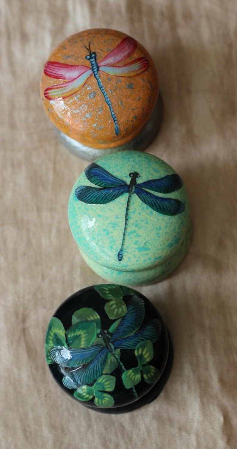 Może przedstawiać: Trzy okrągłe drewniane ozdoby z malowanymi wzorami ważek. G&oacute;rna ozdoba jest pomarańczowa z czerwoną i białą ważką. Środkowa ozdoba jest zielona z niebieską ważką. Dolna ozdoba jest czarna z zieloną i niebieską ważką.