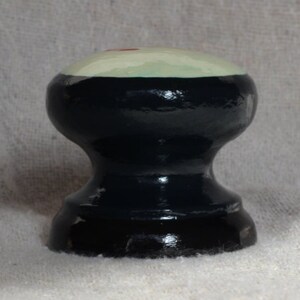Puede incluir: Un pomo decorativo de madera pintado a mano con una parte superior redondeada. La parte superior es de color verde claro con un peque&ntilde;o acento rojo, y el cuerpo es negro. El pomo tiene una forma cl&aacute;sica torneada, adecuada para muebles o decoraci&oacute;n.