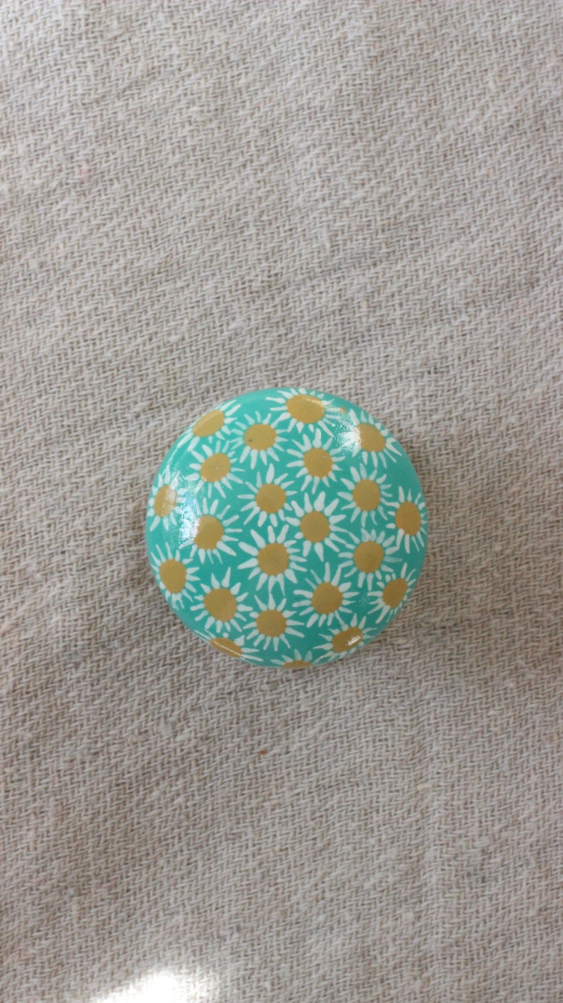 Pode incluir: Uma pedra redonda de cor turquesa pintada com um padr&atilde;o de margaritas brancas e amarelas.