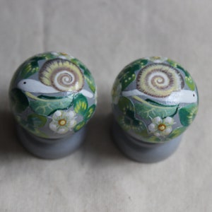 May include: Two decorative, round knobs with a painted snail design. The knobs are primarily gray, with green leaves, white flowers, and a snail with a tan shell. The knobs are mounted on gray bases.