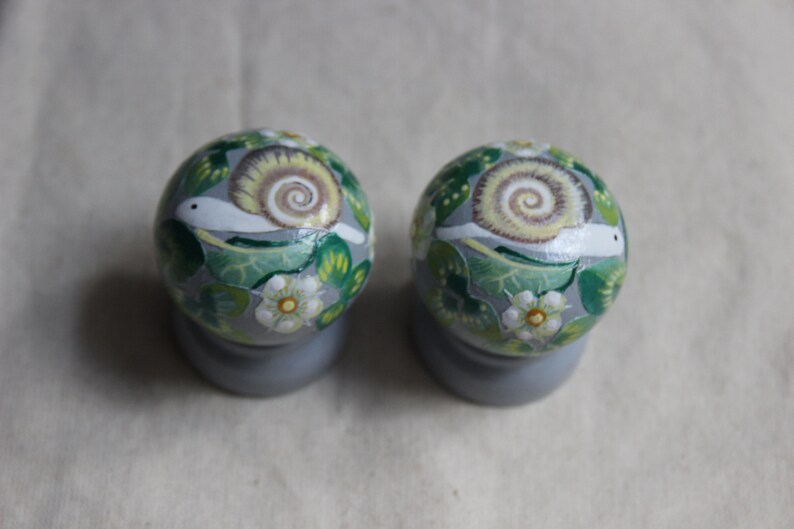 May include: Two decorative, round knobs with a painted snail design. The knobs are primarily gray, with green leaves, white flowers, and a snail with a tan shell. The knobs are mounted on gray bases.