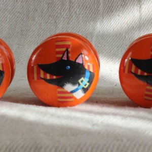 May include: Orange cabinet knobs featuring a painted dog design. The dog has black fur, a white chest, and a blue collar. The background has a cross pattern with red and yellow stripes. These knobs are a decorative hardware item.