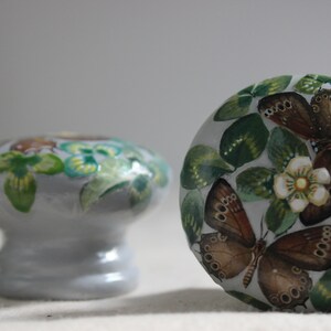 Puede incluir: Un juego de tres pomos de caj&oacute;n de cer&aacute;mica con fondo blanco y dise&ntilde;os pintados de flores y mariposas. Las mariposas son marrones con marcas negras y las flores son blancas con centros amarillos. Las hojas son verdes.