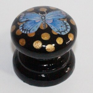 Pode incluir: Um bot&atilde;o de madeira preto com uma borboleta azul e bolinhas douradas.