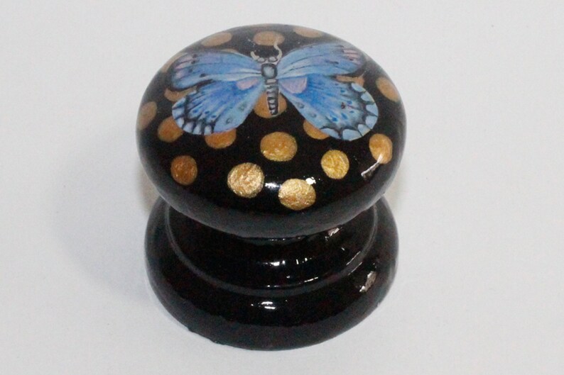 Pode incluir: Um bot&atilde;o de madeira preto com uma borboleta azul e bolinhas douradas.