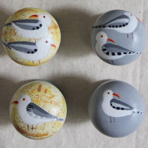 May include: Four round wooden ornaments with painted seagull designs. Two ornaments have a yellow background with white seagulls and a black and grey pattern. Two ornaments have a grey background with white seagulls and a black and grey pattern.