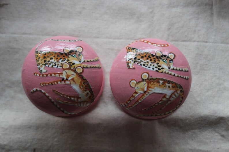 Peut inclure: Deux ornements ronds roses avec des images peintes de l&eacute;opards. Les l&eacute;opards sont dans diff&eacute;rentes poses, certains sont enroul&eacute;s et d'autres sont allong&eacute;s.